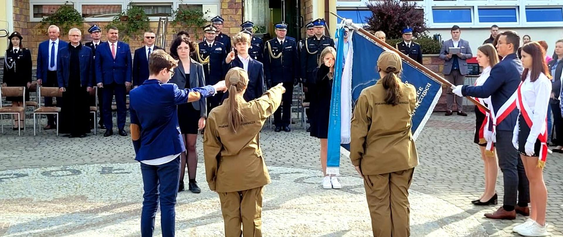 Zdjęcie zrobione na zewnątrz, podczas uroczystego ślubowania uczniów liceum u profilu pożarniczo-ratowniczym. Uczniowie liceum składają ślubowanie wyciągając dłonie w stronę sztandaru szkoły. Za nimi widać zaproszonych gości, wśród nich oficerów Państwowej Straży Pożarnej oraz druhów ochotników.