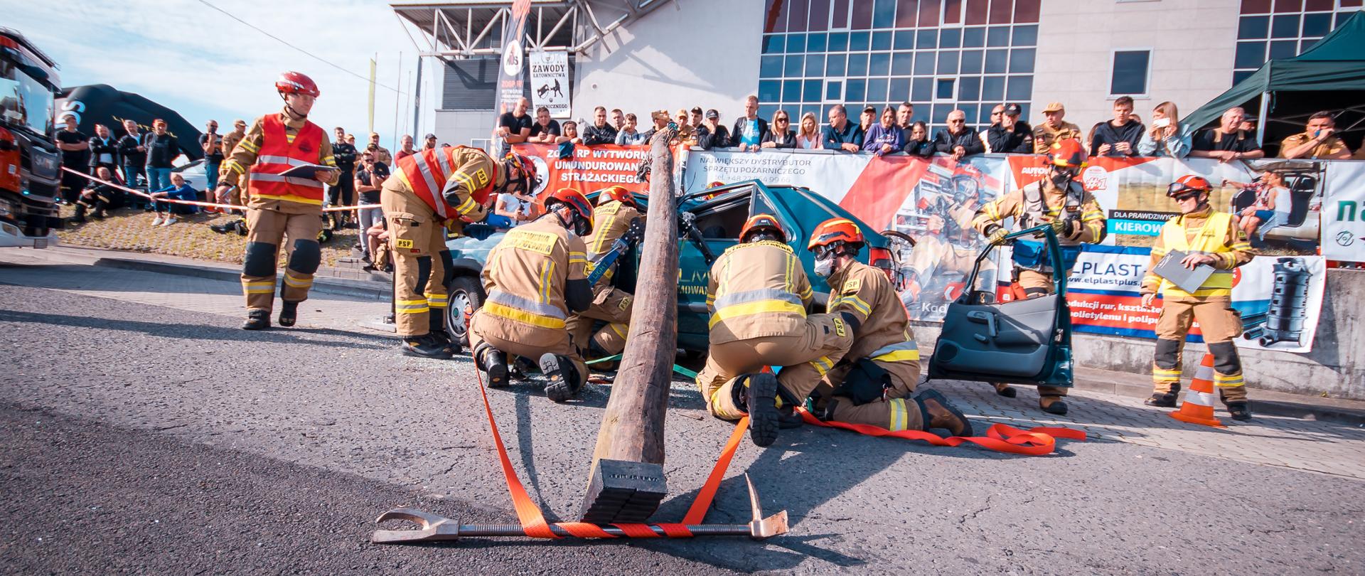 Podczas zawodów strażacy prowadzą działania przy samochodzie przygniecionym drzewem, w tle obserwatorzy 