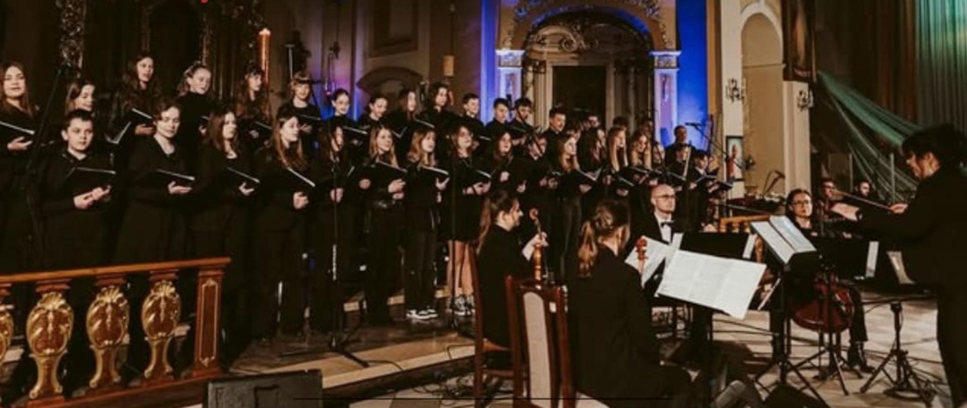 Plakat na jasnoszarym tle. Od góry zaproszenie na „Wspólne Kolędowanie”, poniżej zdjęcie chóru, kwartetu smyczkowego i zespołu z jednego z koncertów, jeszcze niżej na szerokim beżowym pasku szczegóły dotyczące daty, godziny i miejsca wydarzenia, a na samym dole na wąskim szarym pasku informacja o zbiórce charytatywnej z grafiką przedstawiając czerwone serce w objęciach rąk oraz patronat medialny.