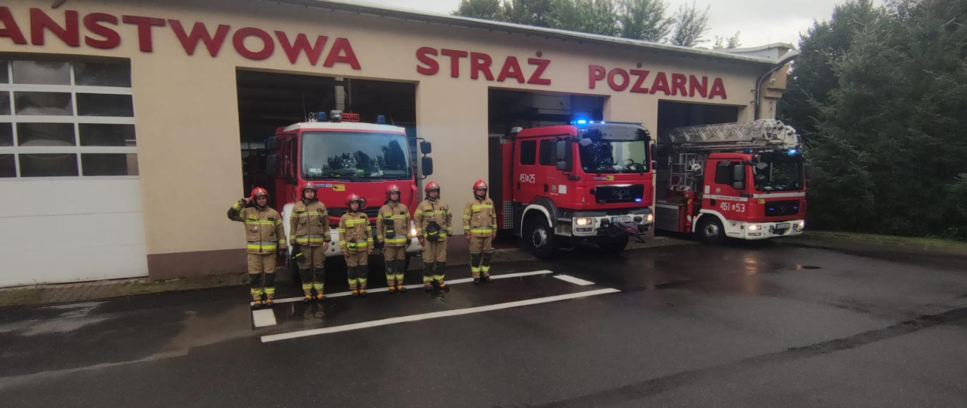 Strażacy oddają hołd w rocznicę Powstania Warszawskiego. Zmiana służbowa, sześciu funkcjonariuszy w ubraniach specjalnych koloru piaskowego ustawieni przed wozami bojowymi oddaje hołd. Z tyłu dostrzegamy trzy stojące pojazdy strażackie z włączonymi światłami.