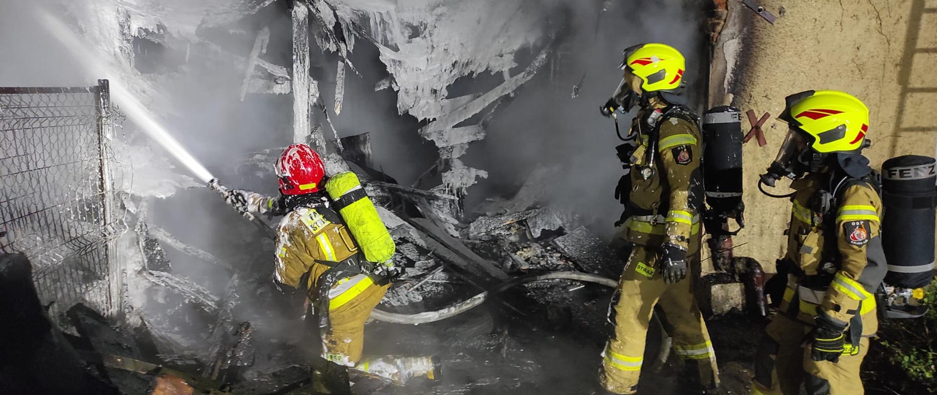Na zdjęciu widzimy trzech strażaków biorących udział w akcji gaszenia pożaru. Strażacy ubrani są w ubrania specjalne koloru piaskowego, na głowie czerwone hełmy, na twarzach maski ochrony dróg oddechowych, na plecach butle z powietrzem. Jeden ze strażaków trzyma prądownice o polewa białą pianą częściowo spaloną konstrukcję. 