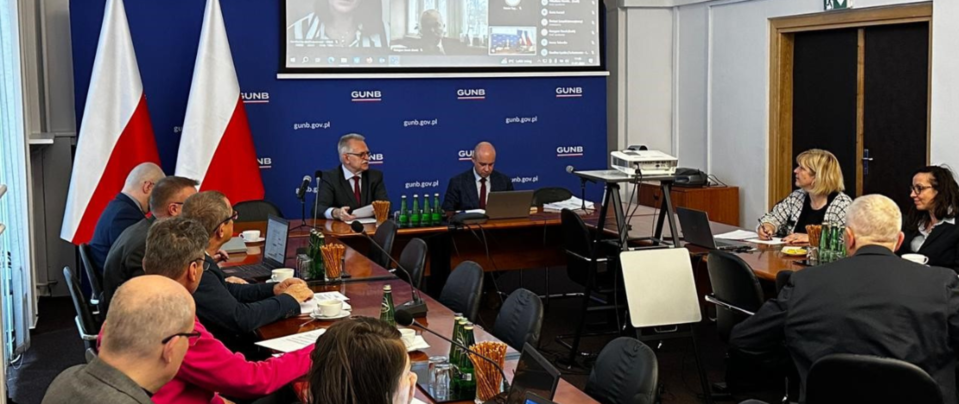 Posiedzenie Rady Wyrobów Budowlanych w sali konferencyjnej z uczestnikami siedzącymi przy dużym stole, flagami Polski w tle oraz ekranem wyświetlającym wideokonferencję.