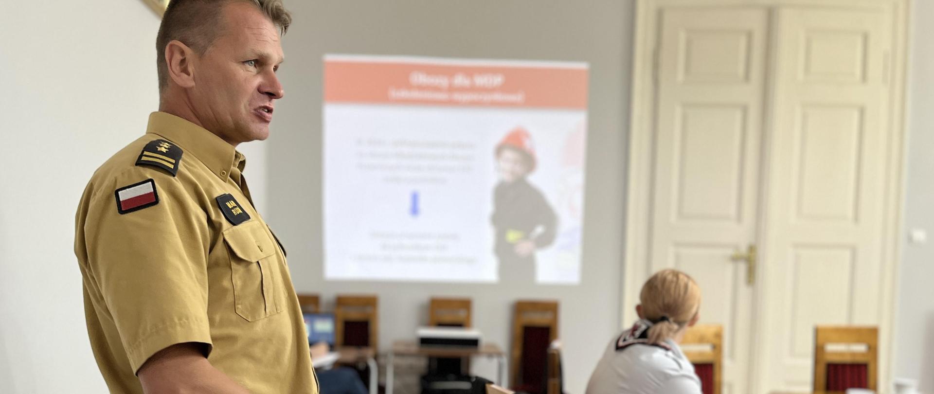 Strażak, w mundurze służbowym stoi przed stołem i prezentuje slajdy. Przed nim widać dwoje innych uczestników spotkania. W tle, na ścianie wyświetlany slajd ze strażakiem.