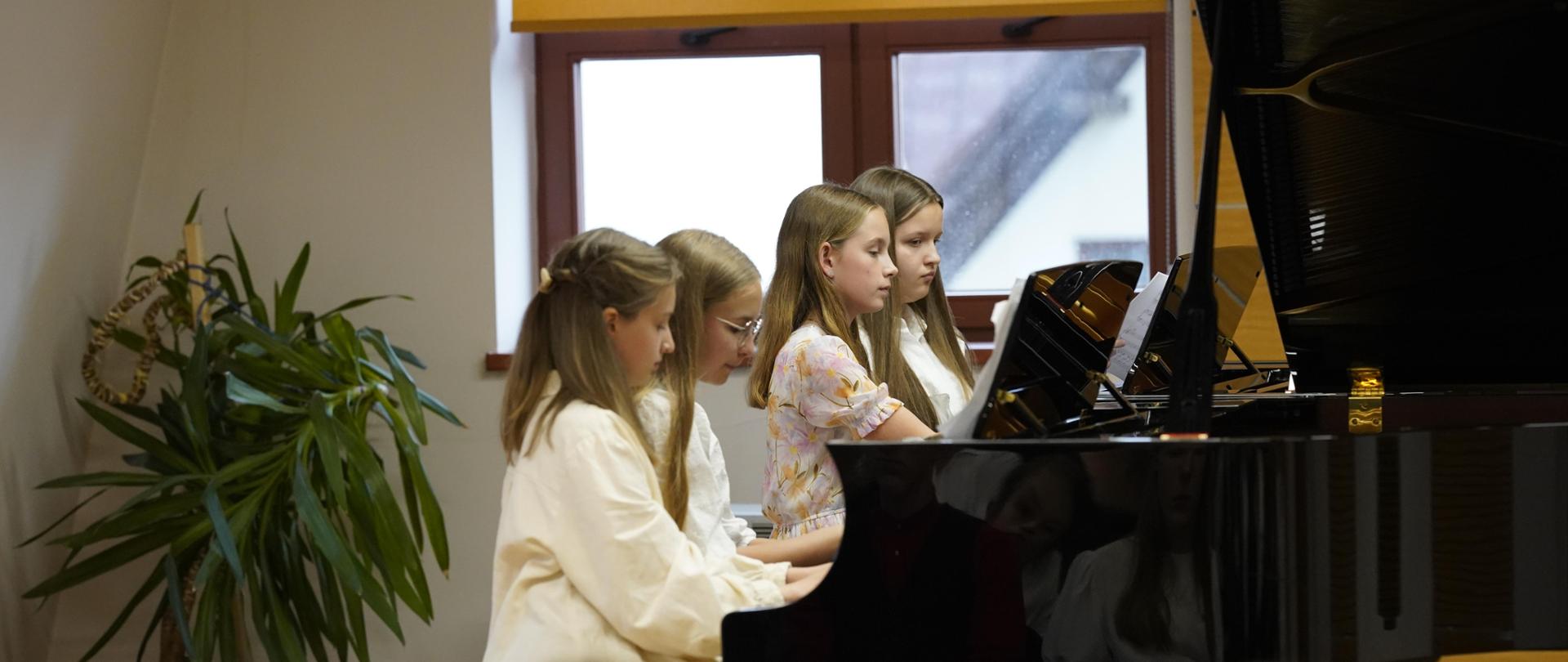 Cztery uczennice grają przy dwóch instrumentach. Na pierwszym planie publiczność. Za uczennicami okno przysłonięte roletą. W rogu sali palma doniczkowa.