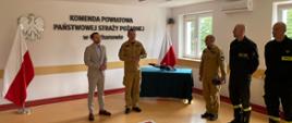 W sali Komendy Powiatowej Państwowej Straży Pożarnej w Ciechanowie, na tle ściany z godłem Polski i napisem instytucji, stoi Komendant Powiatowy PSP w Ciechanowie oraz Prezydent Ciechanowa. Obok nich, przy stole z nożycami hydraulicznymi stoją kolejni strażacy – dwóch w czarnych mundurach bojowych i jeden w piaskowym. W tle widoczne są okna i dwie polskie flagi.