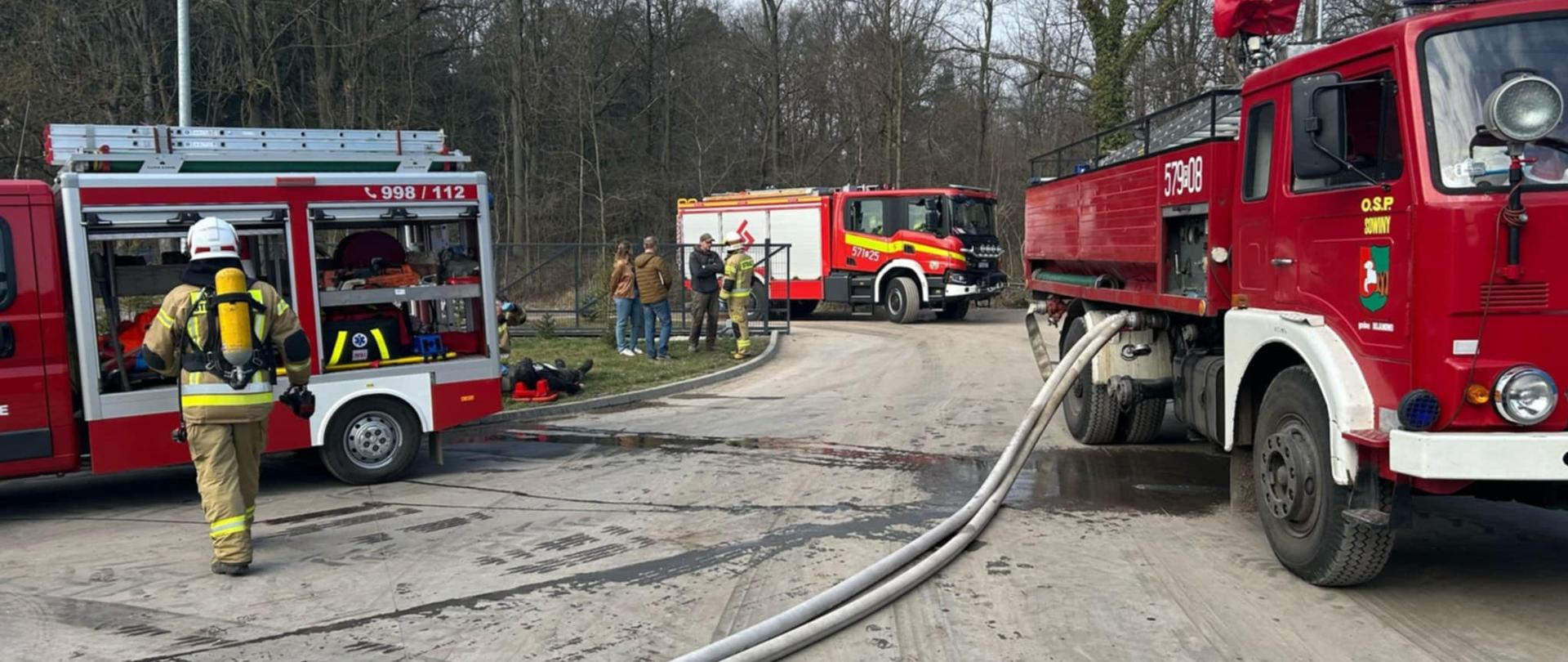 Na terenie zakładu stoją dwa wozy strażackie. Od ciężkiego wozu stojącego z prawej strony rozwinięte są dwie linie wężowe. Przy lekkim samochodzie stojącym z lewej strony, znajduje się strażak mający na sobie aparat ochrony dróg oddechowych. Przed brama wjazdową stoi kolejny pojazd pożarniczy. 