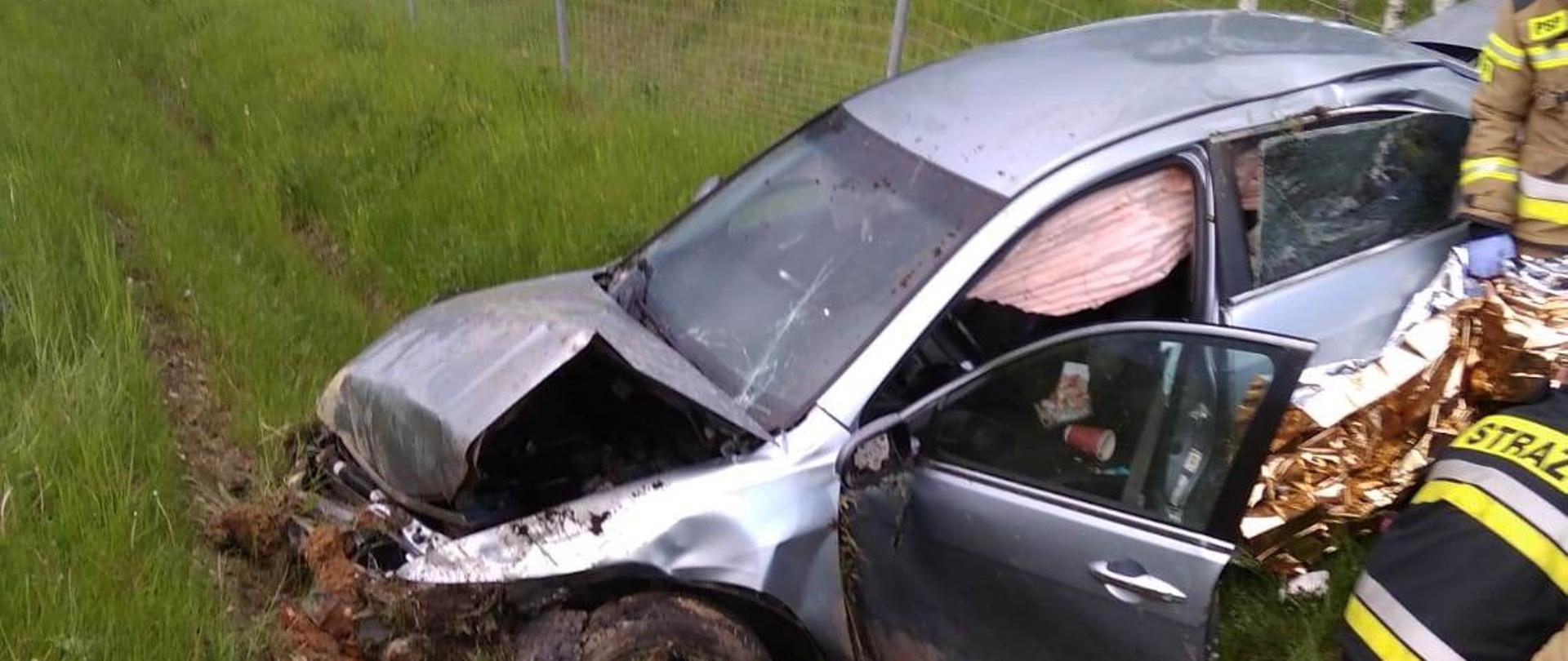 Zdjęcie przedstawia rozbity samochód oraz strażaków PSP udzielających KPP poszkodowanej