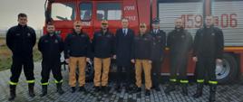 na zdjęciu komendanci psp oraz druhowie OSP pozują do zdjęcia na tle samochodu gaśniczego