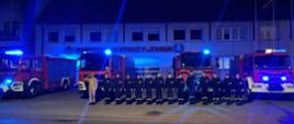 Strażacy oddający hołd dla zmarłego strażaka na tle samochodów pożarniczych z włączonymi sygnałami świetlnymi
