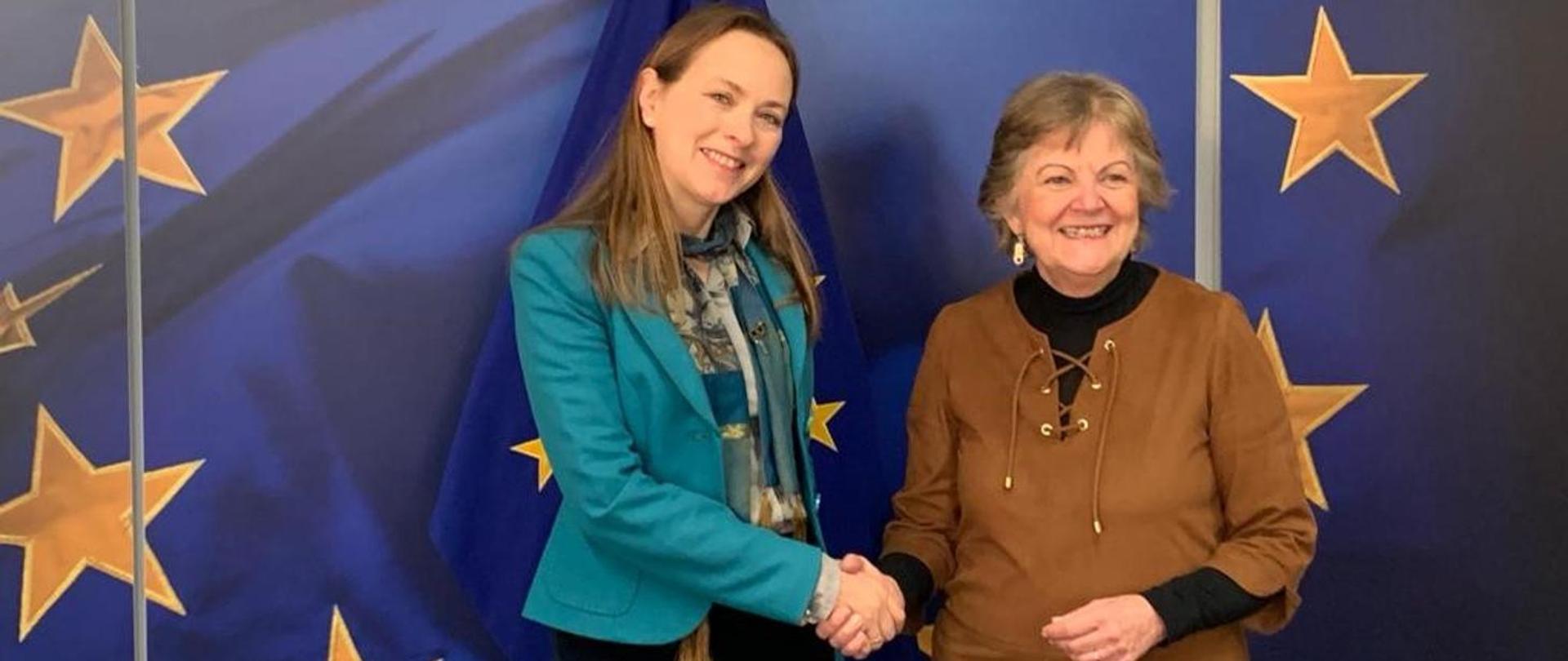 Minister Katarzyna Pełczyńska-Nałęcz i komisarz Elisa Ferreira stoją obok siebie i ściskają sobie dłonie. W tle flaga Unii Europejskiej.