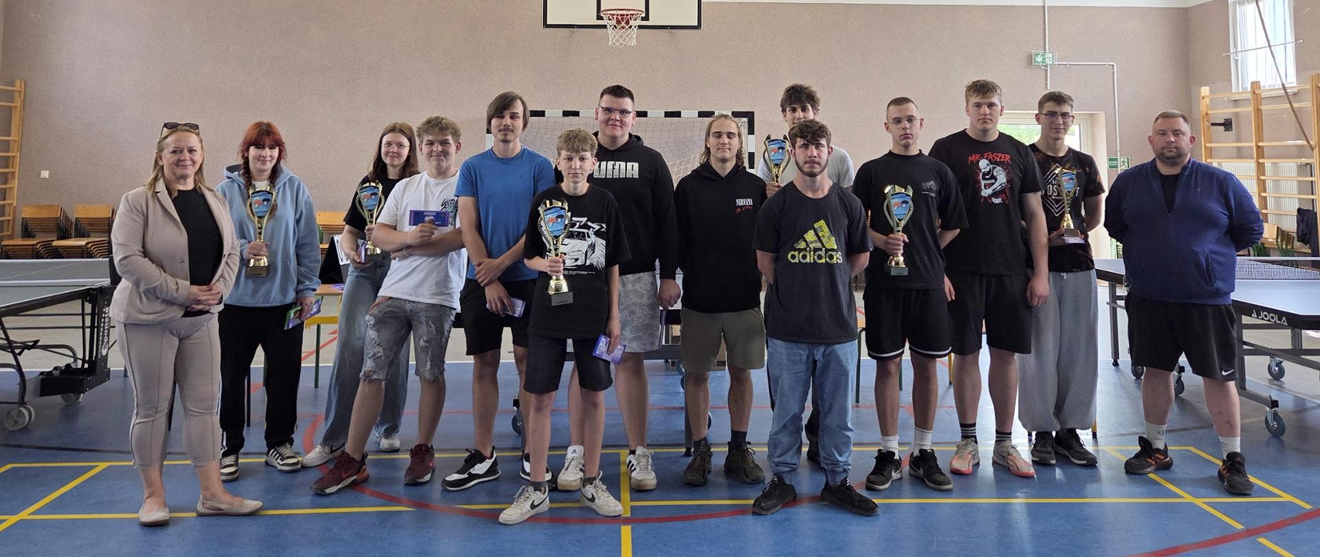 Grupa zawodników i dwoje nauczycieli w sali gimnastycznej.