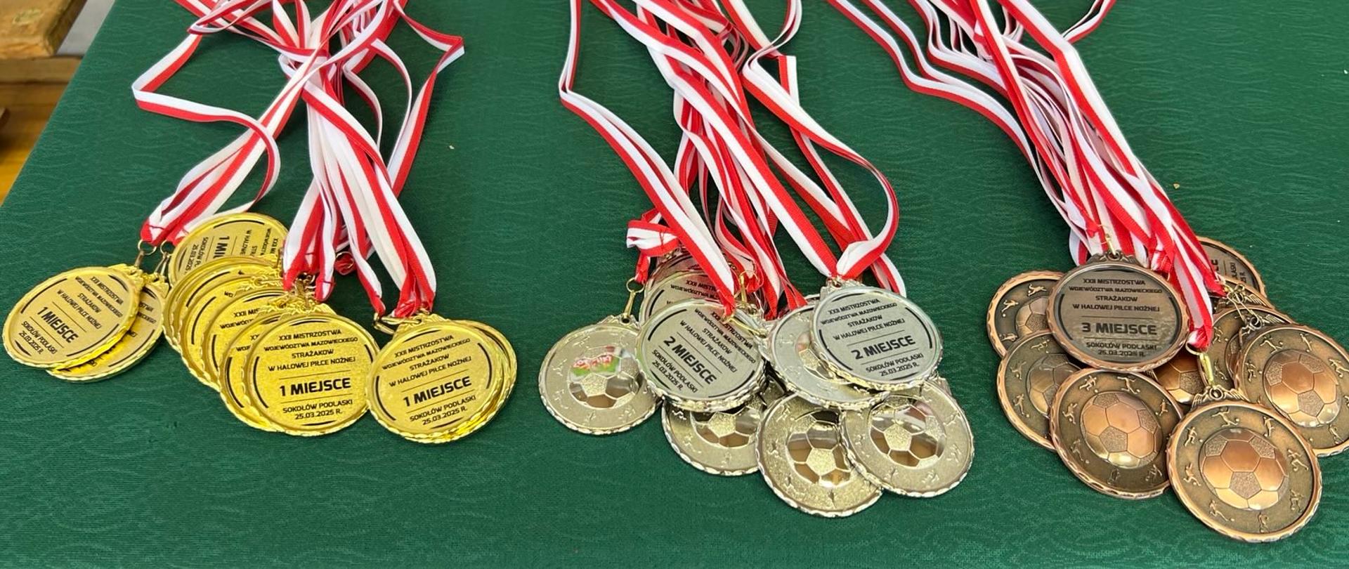 Zdjęcie przedstawia trzy rzędy medali, ułożone na zielonym stole. W każdym rzędzie znajdują się medale w tym samym kolorze. Od lewej są to medale złote, w środku srebrne, a po prawej brązowe. Do każdego medalu doczepiona jest biało-czerwona wstążka.