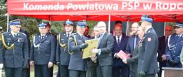 Samorządowcy przekazują na ręce st. bryg. Łukasza Szopy stosowne pamiątkowe adresy, w tle zaproszeni goście
