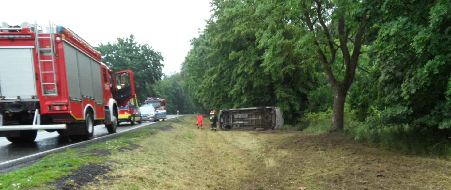wyorane bruzdy w poboczu dalej widać podwozie przewróconego na lewy bok samochodu dostawczego,z lewej strony na drodze samochód pożarniczy, karetka pogotowia, radiowóz i drugi samochód pożarniczy.