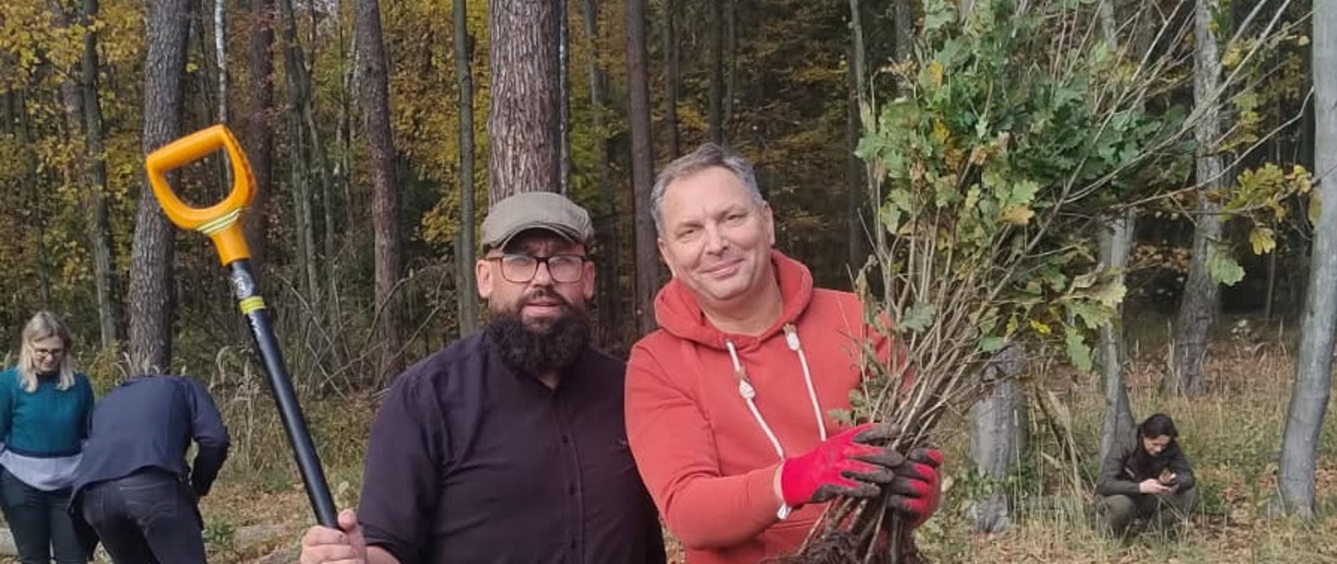 Dwóch meżczyznas stoi w lesie podczas akcji sadzenia lasu. Męzczyzna po lewej stronie trzyma łopatę z pomarańczową rączką, męzczyzna po prawej stronie-ubrany w pomarańczową bluzę -trzyma wiązek sadzonek z dużymi korzeniami. W tle widać kilka osób pracujących przy ziemi oraz drzewa w jesiennych barwach