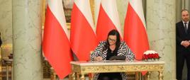Minister Małgorzata Jarosińska-Jedynak składa podpis na akcie powołania na ministra funduszy i polityki regionalnej podczas uroczystości zaprzysiężenia nowego rządu Mateusza Morawieckiego, minister siedzi przy stole i składa podpis, za jej plecami polskie flagi