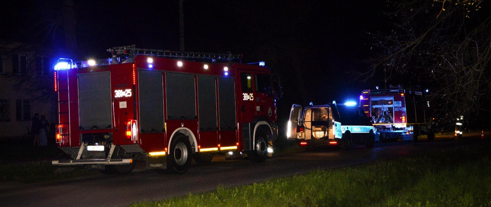 Pora nocna. Na pierwszym planie widoczne samochody służb ratowniczych, które stoją na jezdni jeden za drugim. Za pojazdami, po drugiej stronie jezdni, słabo-widoczny budynek jednopiętrowy. 