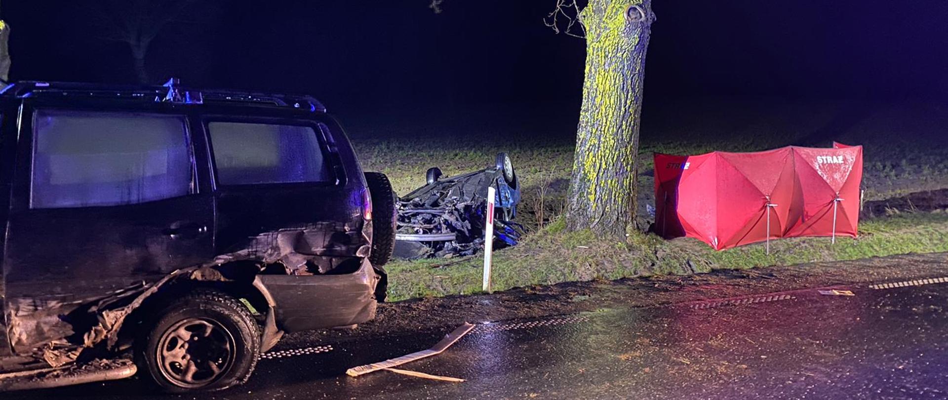 Na zdjęciu znajduje się roztrzaskany samochód z prawej strony oraz z tyłu. Za samochodem znajduje się drzewo obok, którego znajduje się drugi przewrócony samochód, z drugiej strony drzewa rozstawiony jest czerwony parawan z napisem STRAŻ.