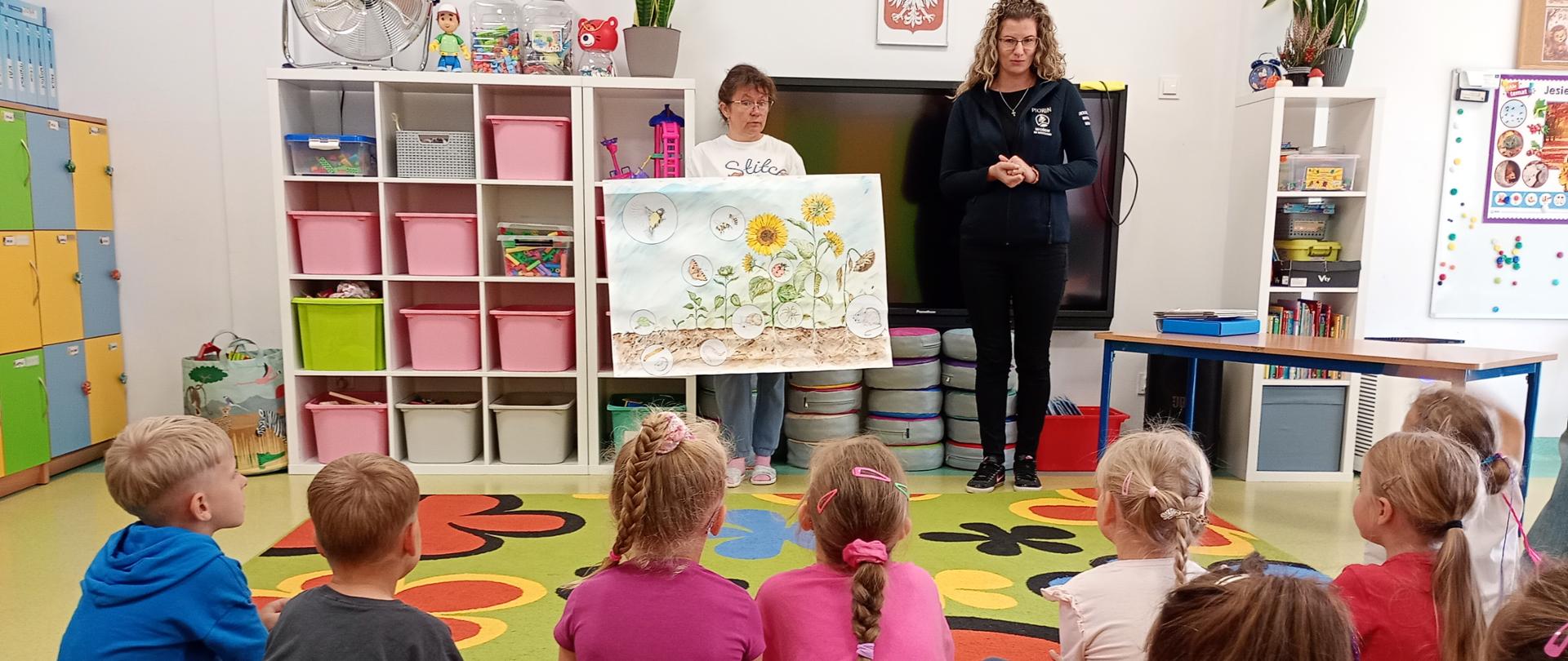Grupa przedszkolaków siedząca w sali przedszkolnej na kolorowym dywanie w kwiaty. Przed nimi stoją dwie kobiety – jedna z nich trzyma dużą planszę edukacyjną ilustrującą rozwój słonecznika, a druga opowiada. W tle widać regały z różowymi i zielonymi pojemnikami oraz szafki.
