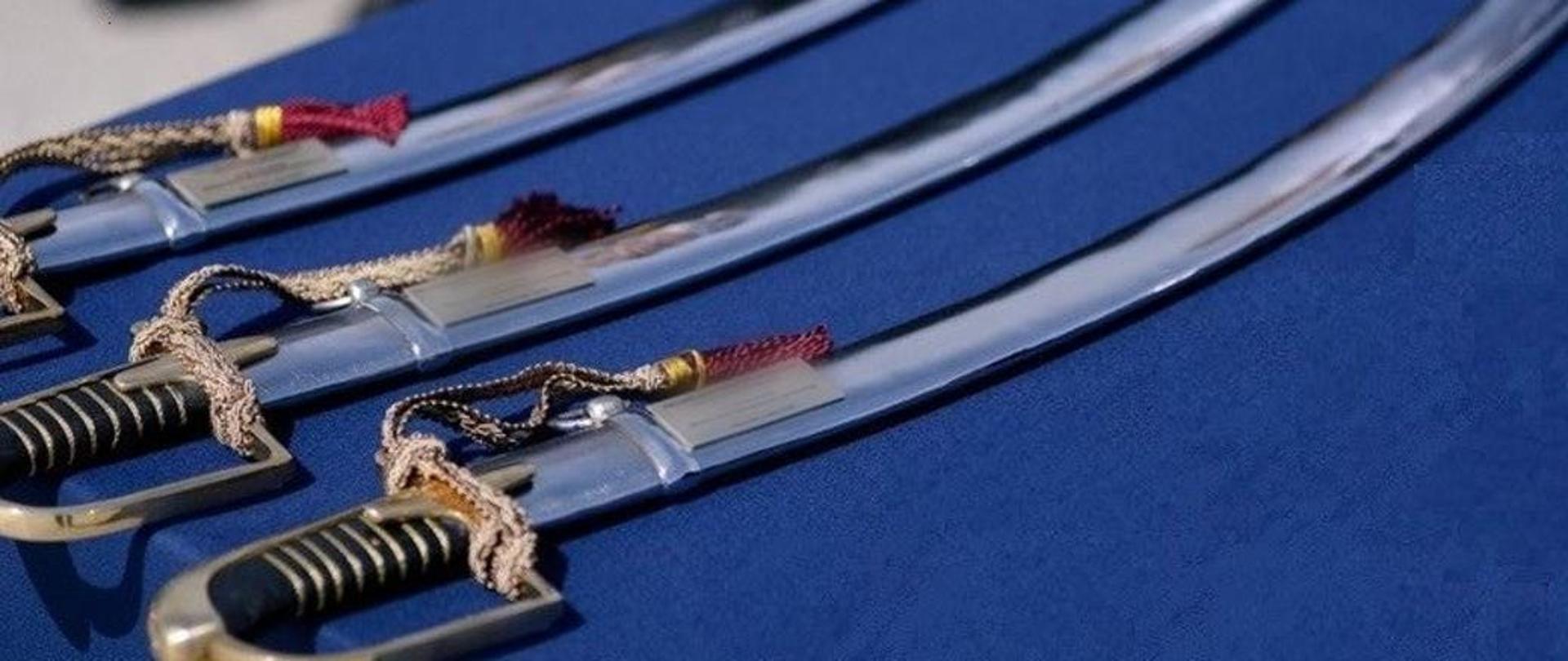 Na zdjęciu widoczne szable. Szable ułożone jedna obok drugiej. Leżą na niebieskim materiale.