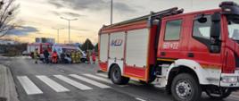 
Na zdjęciu czrwony samochód strażacki, w tyle strażacy wraz z ratownikami medycznymi udzielający pomocy poszkodowanemu, za nimi wrak pojazdu oraz kasretka i samochód strażacki