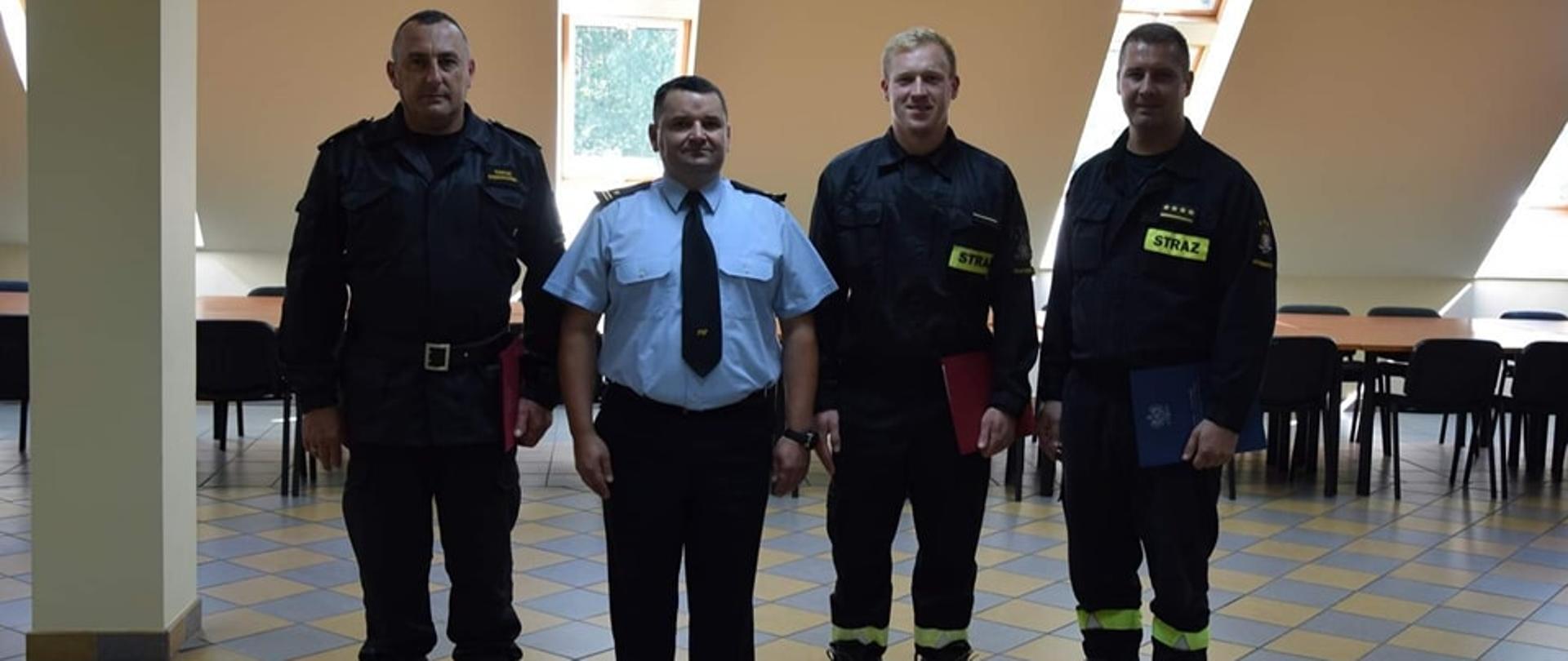 Na zdjęciu widać czterech mężczyzn- strażaków ustawionych w szeregu. W niebieskiej koszuli stoi Komendant Powiatowy młodszy brygadier Witold Juszczak, po jego lewej i prawej stronie stoją strażacy trzymający w dłoniach teczki.