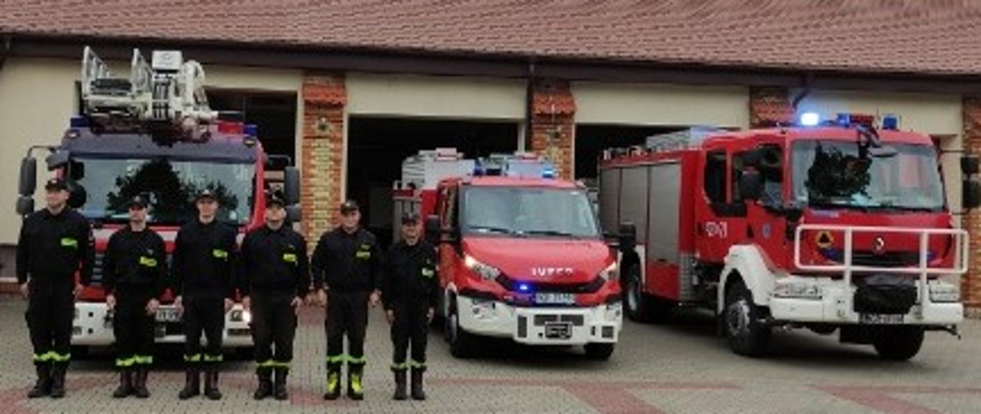 Strażacy PSP stoją w rzędzie przed pojazdami wyprowadzonymi przed bramy garażowe.