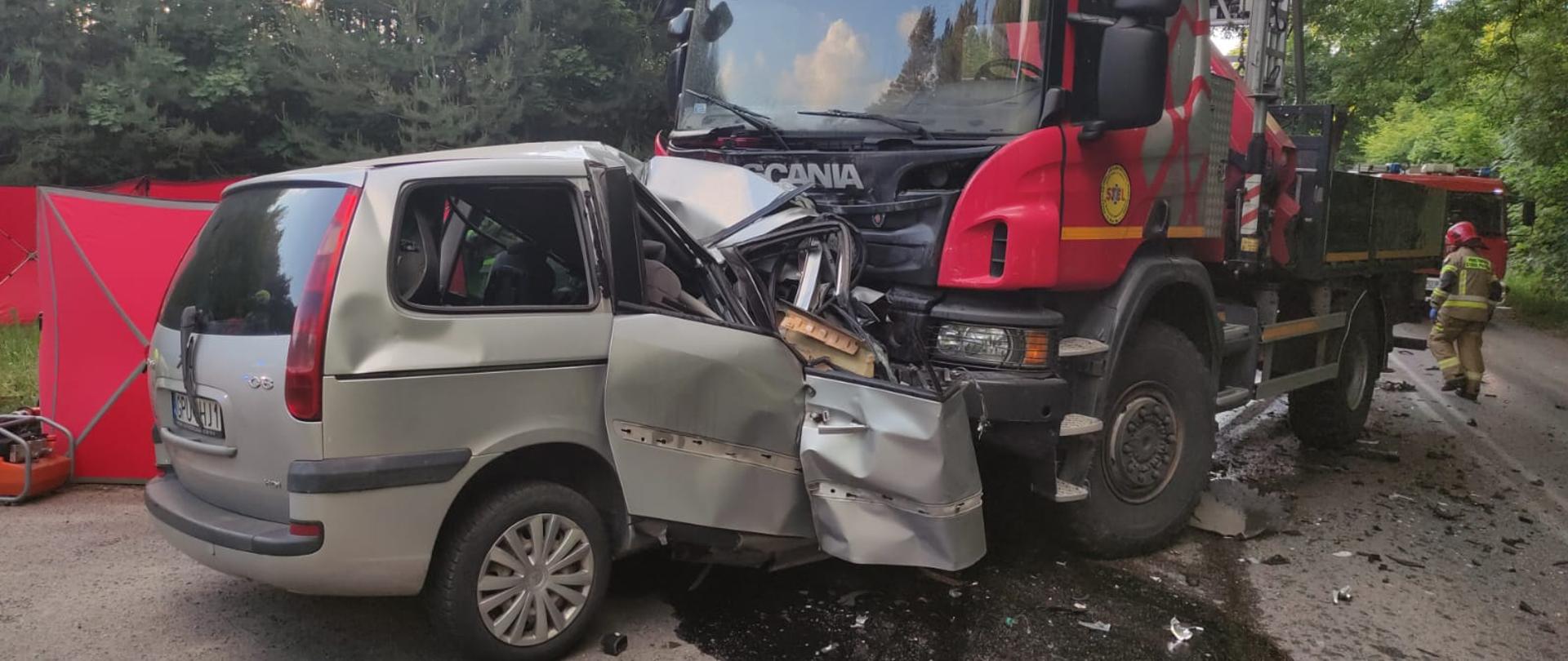 Na zdjęciu widoczny samochód osobowy (minivan) koloru srebrnego, który wbił się pod samochód ciężarowy. Przód samochody osobowego cały rozbity.. Spod samochodu wylały się płyny eksploatacyjne.