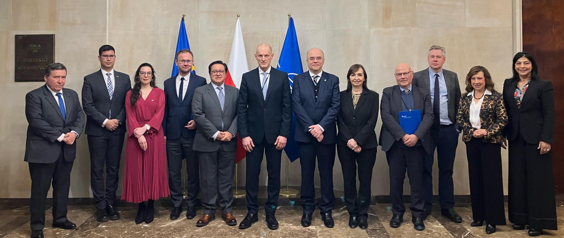 Grupa osób w formalnych strojach pozuje do wspólnego zdjęcia w reprezentacyjnej sali. W tle widoczne są flagi oraz marmurowa ściana, a nad nimi ozdobny żyrandol. Uczestnicy stoją w jednym rzędzie, patrząc w stronę aparatu.