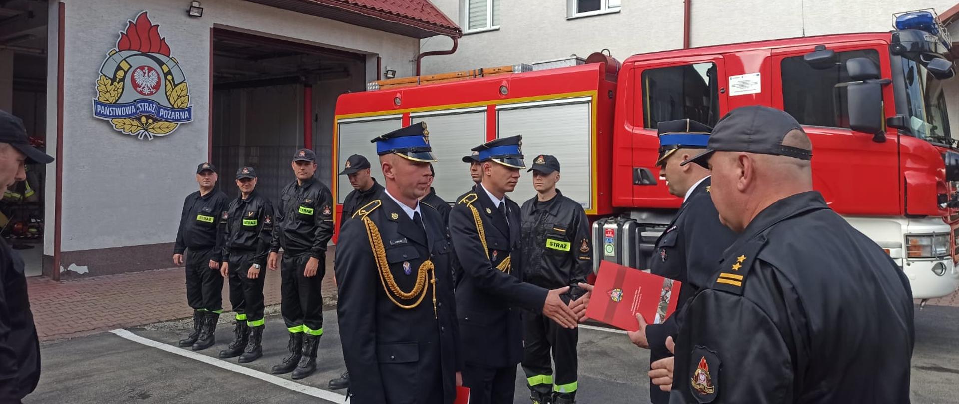Na zdjęciu widać dwóch funkcjonariuszy PSP oraz komendanta w mundurach galowych oraz zastępce komendanta w ubraniu dowódczo-sztabowym na placu przed komendą PSP w Ustrzykach Dolnych. Komendant wraz z zastępcą witają nowych funkcjonariuszy w szeregach komendy. W tle widać zmianę służbową, wóz bojowy oraz budynek komendy PSP w ustrzykach Dolnych.