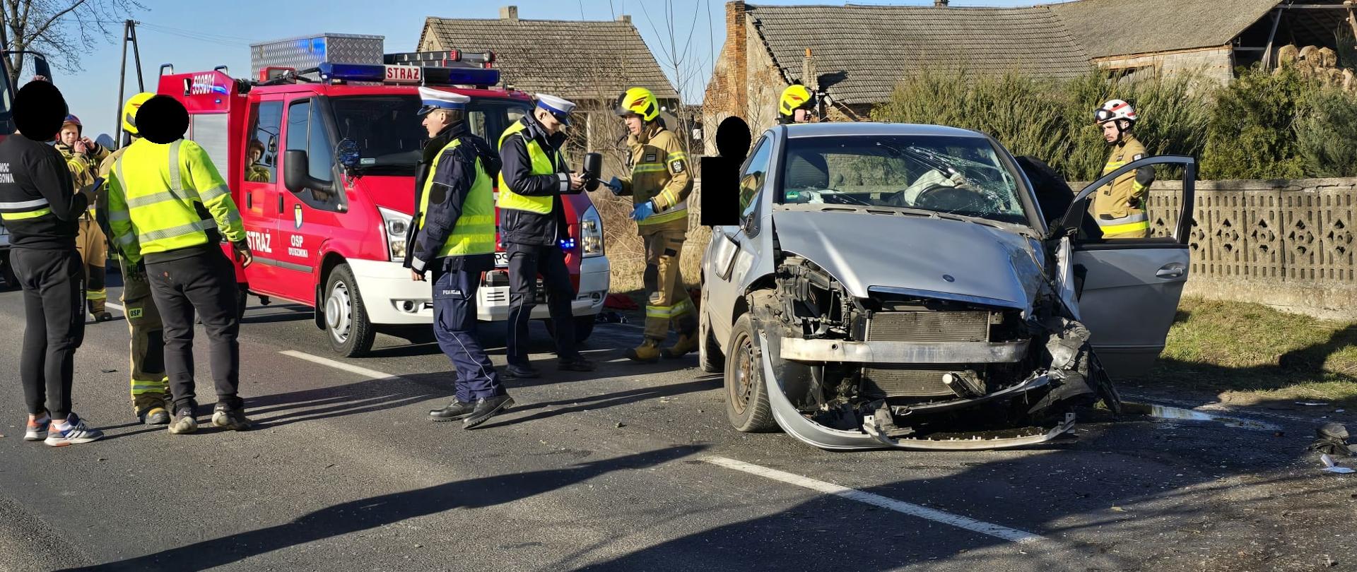 Uszkodzony samochód na drodze, za nim pojazd straży, w pobliżu osoby