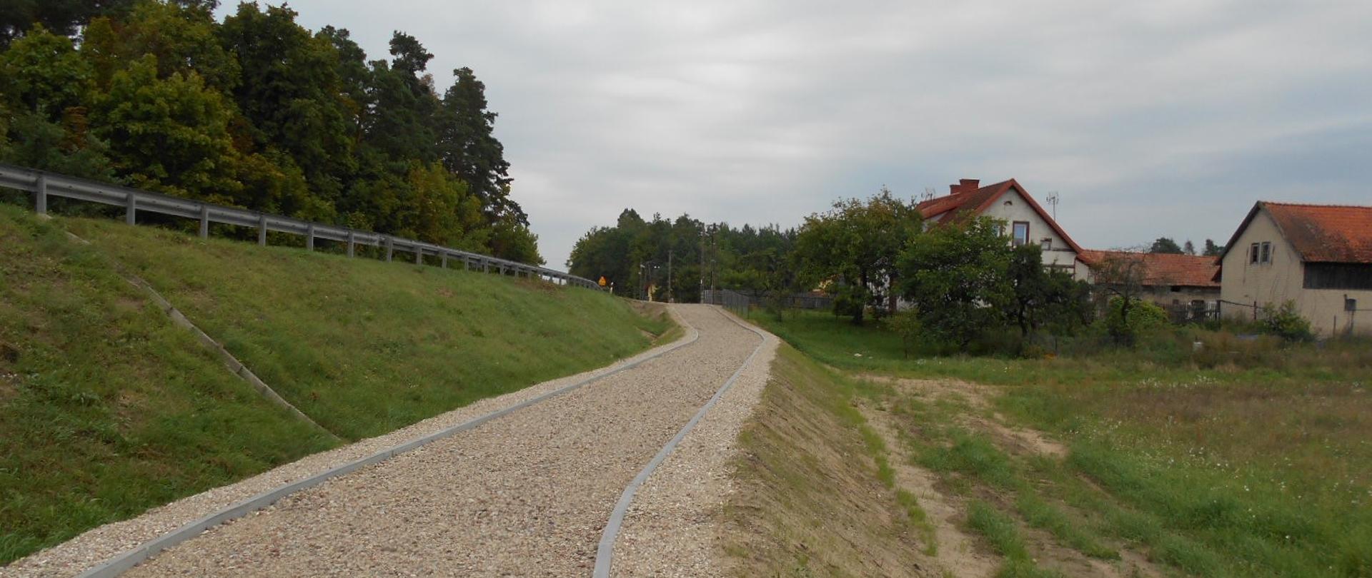 Droga rowerowa o żwirowej nawierzchni, biegnąca wzdłuż drogi na nasypie. Po prawej stronie zabudowania.