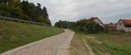 Droga rowerowa o żwirowej nawierzchni, biegnąca wzdłuż drogi na nasypie. Po prawej stronie zabudowania.