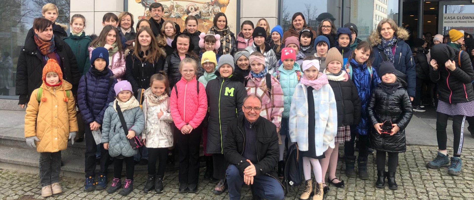 Na zdjęciu grupa uczniów wraz z opiekunami podczas szkolnej wycieczki. Wszyscy w kurtkach, niektórzy maja czapki. Zdjęcie wykonano przed Teatrem Muzycznym w Gdyni.