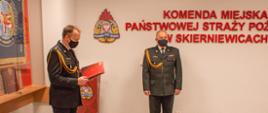 Zdjęcie przedstawia dwóch strażaków komendy miejskiej w mundurach wyjściowych koloru czarnego na tle czerwonego napisu Komenda Miejska Państwowej Straży Pożarnej w Skierniewicach, loga psp. Jeden ze strażaków odczytuje rozkaz personalny.