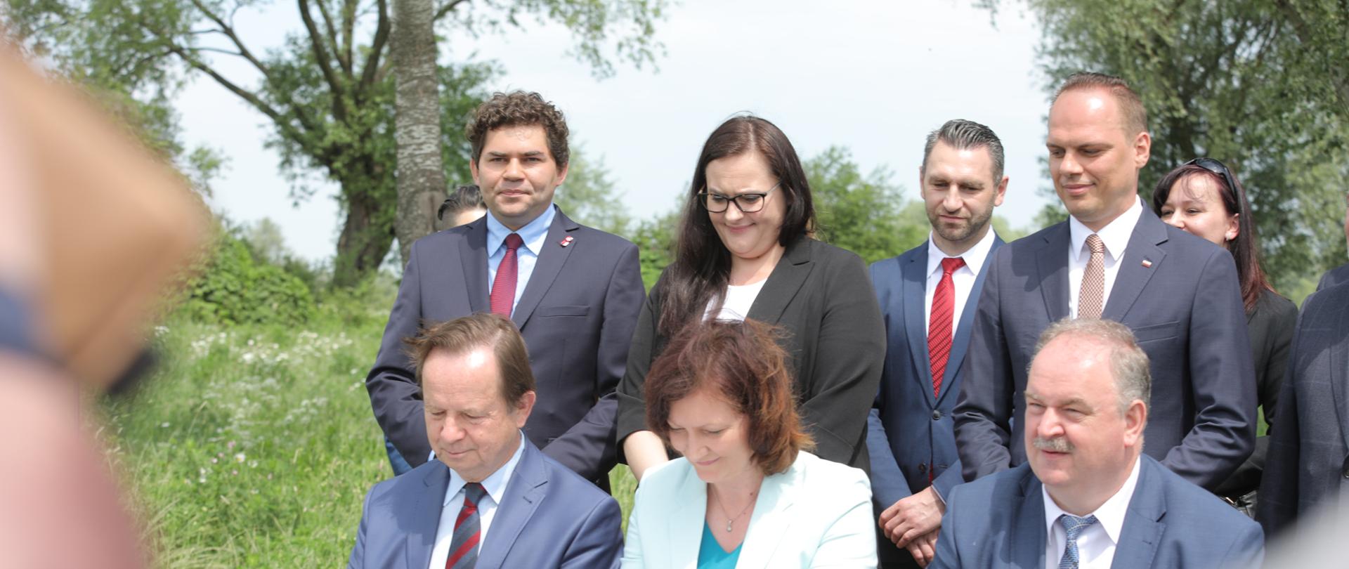 Na zdjęciu widocznych jest siedem osób , trzy z nich siedzą przy stole i podpisują dokumenty. Cztery pozostałe stoją za ich plecami.