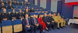 Na sali widowiskowej siedzą zaproszeni goście, strażacy ubrani są w mundury, w tle czerwone podświetlenia paneli