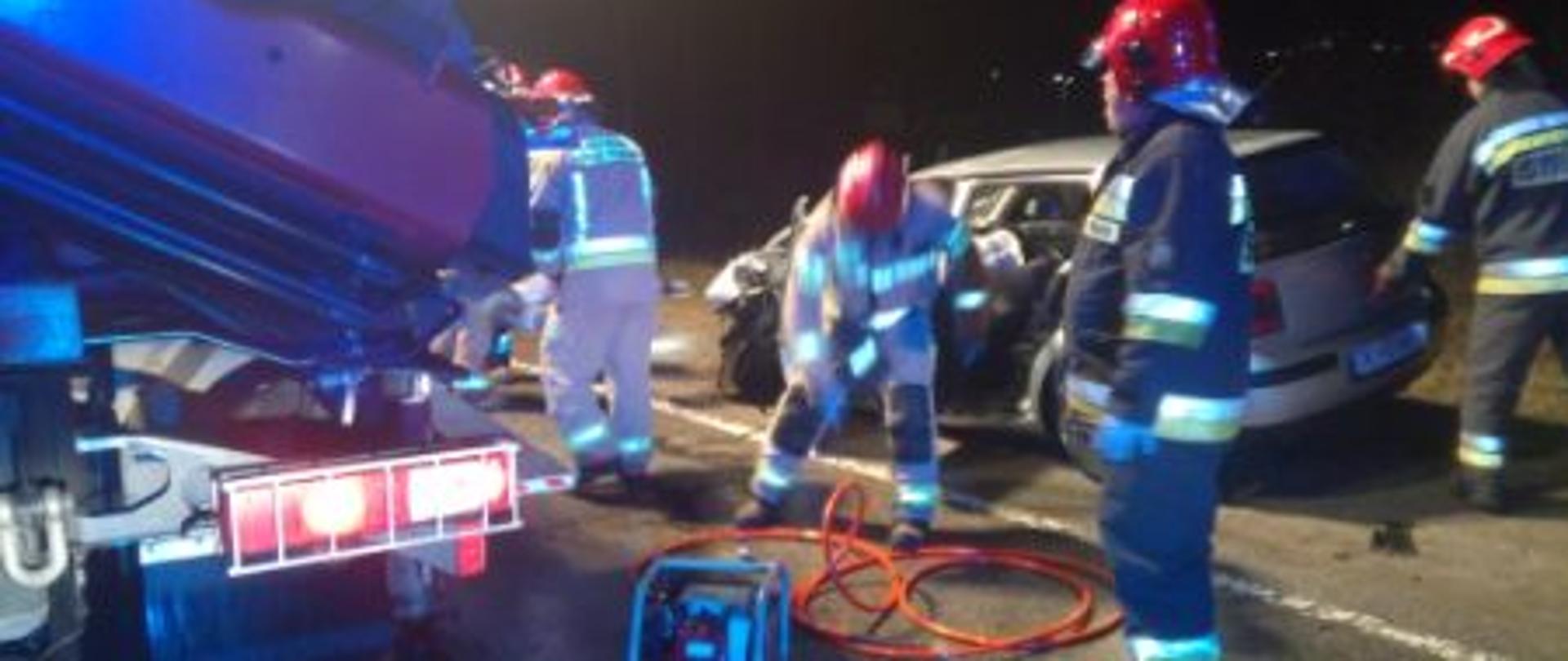 Po lewej stronie widać fragment samochodu pożarniczego ratownictwa technicznego. W centralnej części grupa ratowników podczas akcji z czego jeden pochylony nad wężami hydraulicznymi zasilającymi nożyce. W tle widać zniszczony w wyniku zderzenia samochód osobowy.