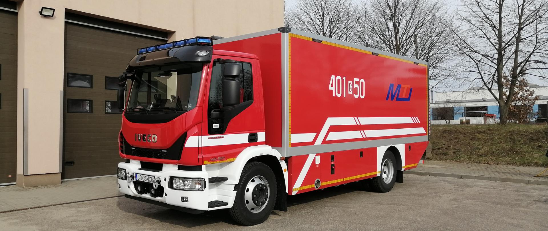Przedstawia samochód strażacki w kolorze czerwonym z dwoma białymi paskami, który ustawiony jest na parkingu, a w tle znajduje garażu