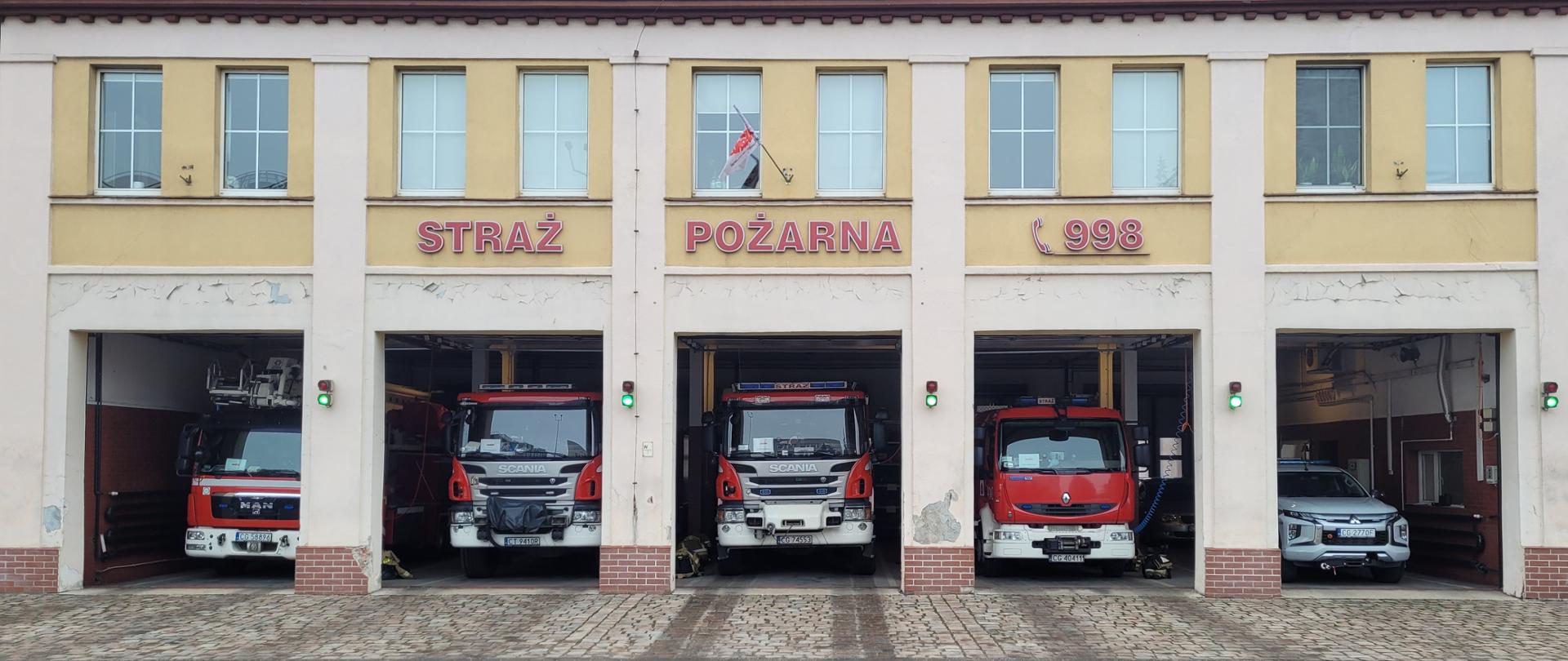 Jasny budynek z pięcioma prostokątnymi otworami wyjazdowymi, w których stoją czerwone i białe strażackie samochody.