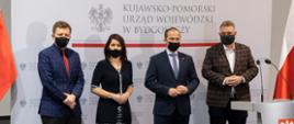 Trzech mężczyzn i kobieta stojących na tle biało-czerwonych flag i logo Urzędu Wojewódzkiego