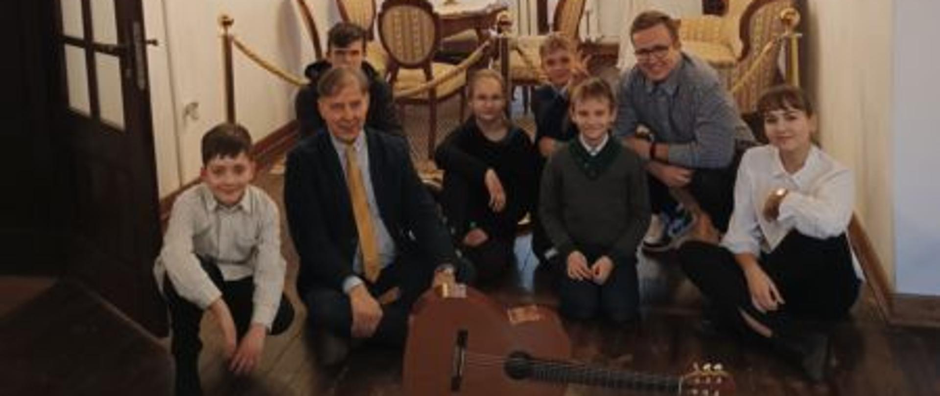 Sześcioro uczniów i nauczyciel siedzą na drewnianej podłodze w salonie u Lutosławskich. Przed nimi położona gitara klasyczna. Za uczniami salonik z meblami z XIX wieku.
