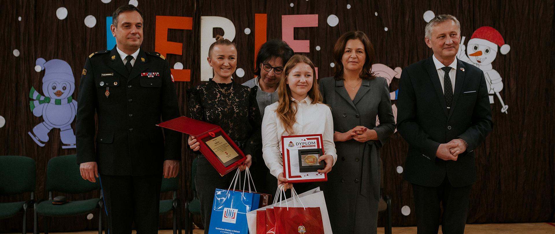 Na zdjęciu widzimy od lewej strażaka w mundurze wyjściowym PSP w stopniu nadbrygadiera, kobietę trzymającą upominki, kobietę w okularach, dziewczynkę trzymającą upominki, kobietę oraz mężczyznę w strojach wizytowych. Wszyscy się uśmiechają.