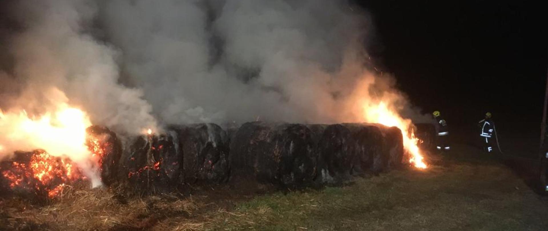 Pożar stogu balotów, nad stogiem gęsty dym po prawej stronie strażacy gaszą pożar.