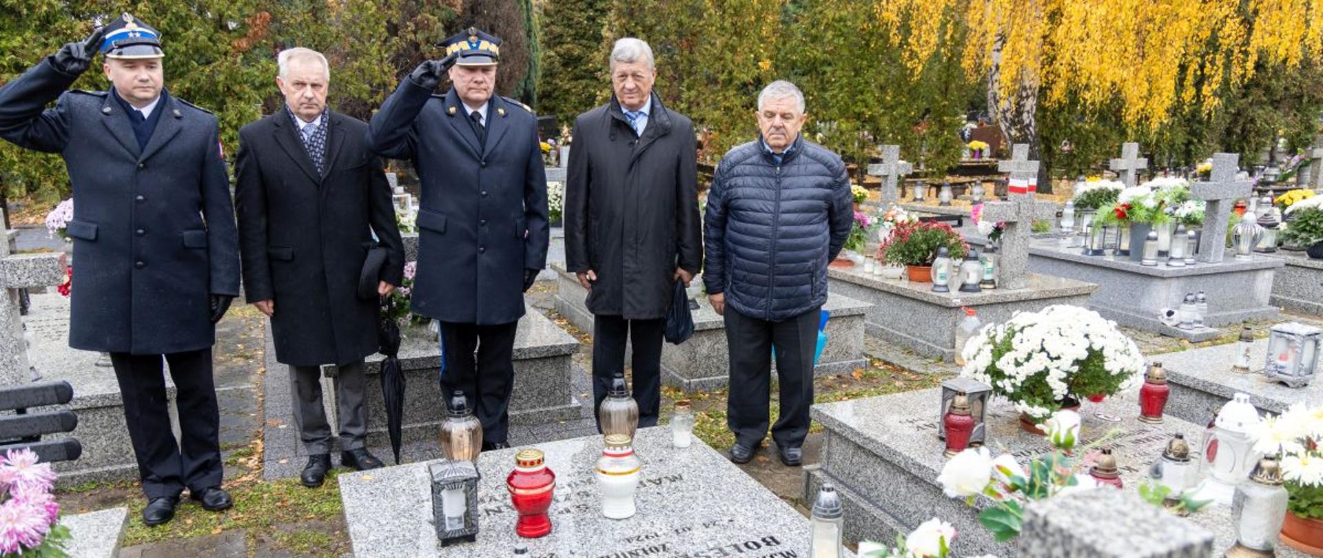 Delegacja PSP, złożona z pięciu mężczyzn w mundurach i cywilnych ubraniach, oddaje hołd zmarłym strażakom przy grobach ozdobionych kwiatami, zniczami i biało-czerwoną wstęgą. Dwóch funkcjonariuszy salutuje, podkreślając ceremonialny charakter wizyty.