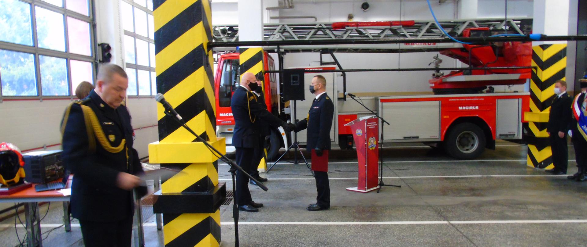 na zdjęciu w garażu widoczny strażak w mundurze wyjściowym prowadzący ceremonię dalej Zastępca Łódzkiego Komendata Wojewódzkiego składający podziękowanie Zastępcy Komendanta Powiatowego Państwowej Straży Pożarnej w Bełchatowie w tle samochód bojowy-drabina