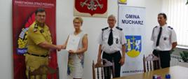 Widok z przodu. Uczestnicy spotkania stoją za stołem na tle godła Polski i rolapów KP PSP Wadowice i Gminy Mucharz. Od lewej: komendant powiatowy PSP, który ściska dłoń wójta gminy, prezes OSP i naczelnik OSP.