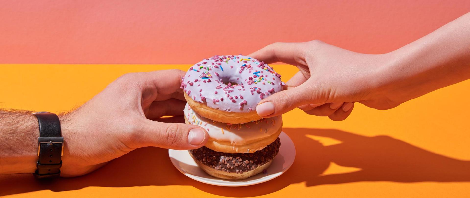 Na zdjęciu są trzy donuty ułożone na talerzu. Z prawej strony widoczna jest damska dłoń a z lewej męska. Obie wysunięte są w stronę pączków. Tło jest pomarańczowo-różowe.