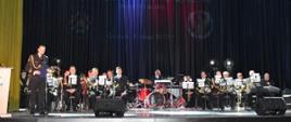 Wojskowy w mundurze wyjściowym ze sznurem przemawia do mikrofonu za nim siedzą żołnierze z instrumentami muzycznymi.