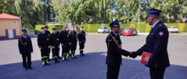 Mł. bryg. Andrzej Wróbel wręcza akt mianowania st. kpt. Daniela Kaczmarka na stanowisko Zastępcy Dowódcy JRG w Oleśnie. Strażacy w mundurach galowych. W tle widoczni funkcjonariusze ubrani w ubranie koszarowe."
